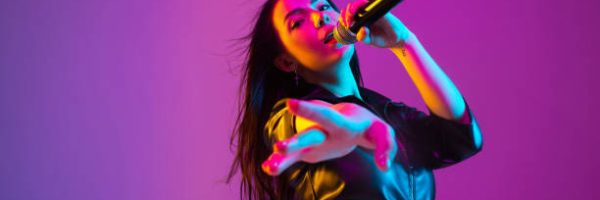 Caucasian female singer portrait isolated on purple studio background in neon light. Beautiful female model in black wear with microphone. Concept of human emotions, facial expression, ad, music, art.