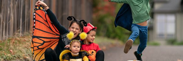 Four mixed race siblings in costumes happily play together outside in their driveway on Halloween. An elementary age boy dressed as a superhero is jumping over a large white pumpkin.